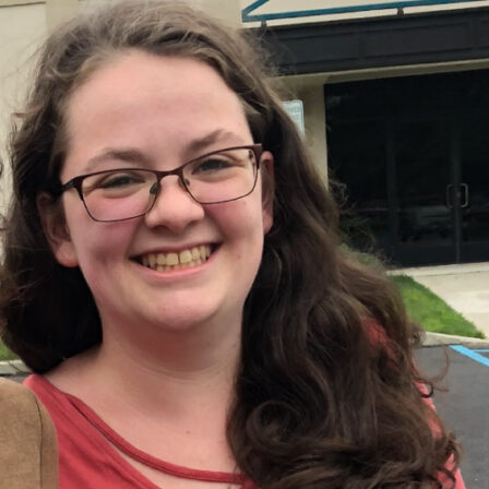 Alyssa Koeck, a smiling young woman with long brown wavy hair, wearing glasses and a red scoopneck shirt, standing on the lawn in front of a home