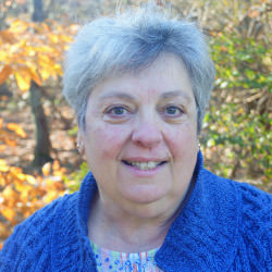 Cindy DeLuca, a smiling woman with short gray hair, wearing a patterned shirt and a royal blue sweater, standing in front of a flower garden planted next to a fence