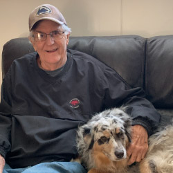 John Barnak, a smiling gray-haired man wearing a tan baseball cap, glasses, and a blue pullover sweatshirt, sitting on a black couch with his arm around a multi-colored dog