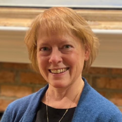 Kelli Pisciotti, a smiling woman with short blond hair and bangs standing against a brick wall, wearing a silver necklace, a black shirt and a blue jacket