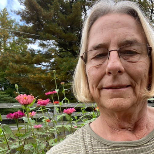 Linda Raudabaugh, a woman with shoulder length blond hair, wearing glasses and a striped green shirt, standing in front of a flower garden planted next to a fence