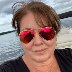 Lisa Cardwell, a smiling woman with short brown hair and bangs sitting on a boat on a lake in front of a shoreline with trees and cloudy skies, wearing sunglasses and a black shirt