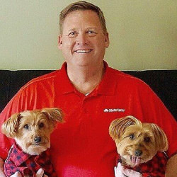 Michael Peterson, a smiling man with short blond hair, wearing a red shirt with a State Farm logo, sitting on a couch holding two small brown dogs