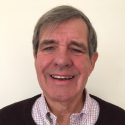 Paul Schmid, a smiling gray-haired man standing against a cream-colored wall, wearing a checkered shirt and dark cardigan sweater