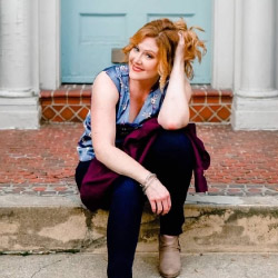 Sarah Wallace, a smiling woman sitting on a brick and cement porch step in front of a blue front door, holding her long wavy strawberry-blond hair up with her hand, wearing a sleeveless blue denim shirt and dark pants