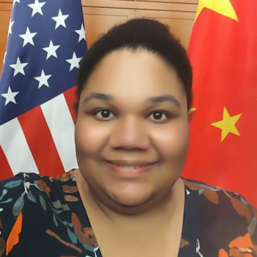 Shayna Canty, a smiling young black woman with natural black hair pulled back into a ponytail, wearing a patterned shirt and standing in front of an American flag and a People’s Republic of China flag