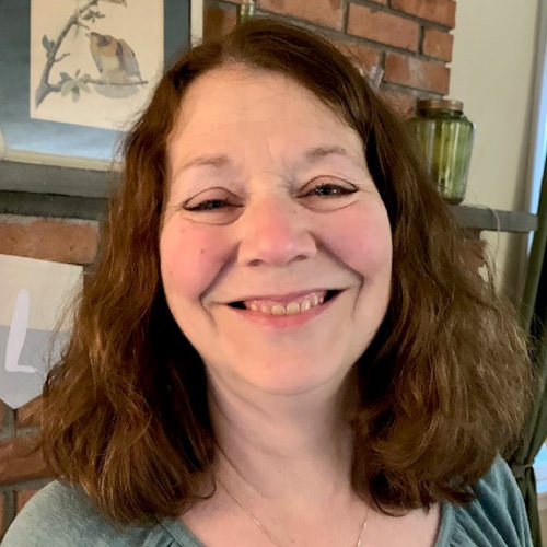 Stacey Koeck, a smiling woman with long brown wavy hair, wearing a silver necklace and standing in front of a brick fireplace mantel