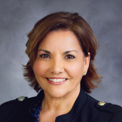 TJ Price, a smiling woman with shoulder length wavy brown hair and brown eyes posed against a textured gray wall, wearing a dark shirt with epaulets