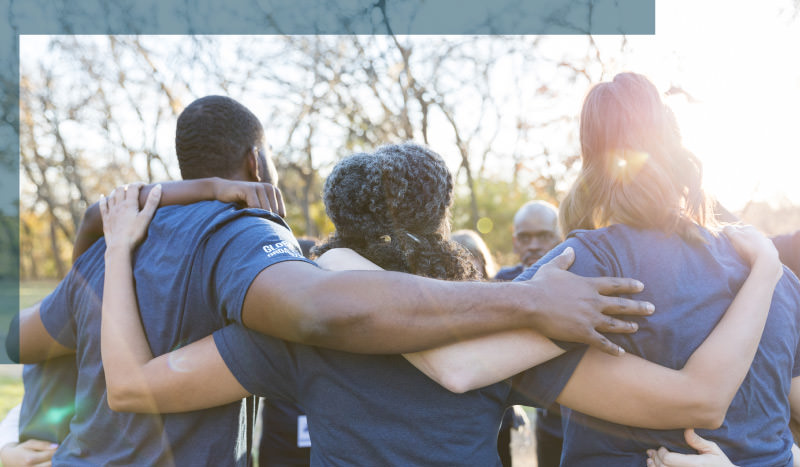 Group of volunteers in navy blue shirts hugging in a circle with trees and sky in the background