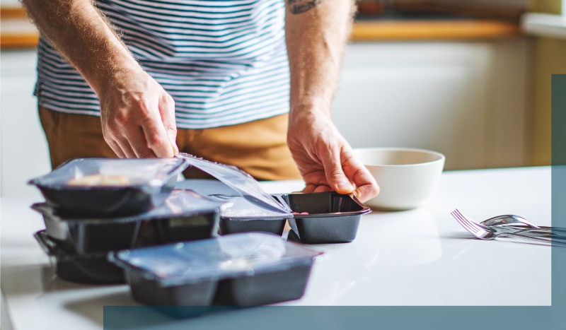 Man opening black container with prepared food, while wearing brown pants and a blue and white striped t-shirt