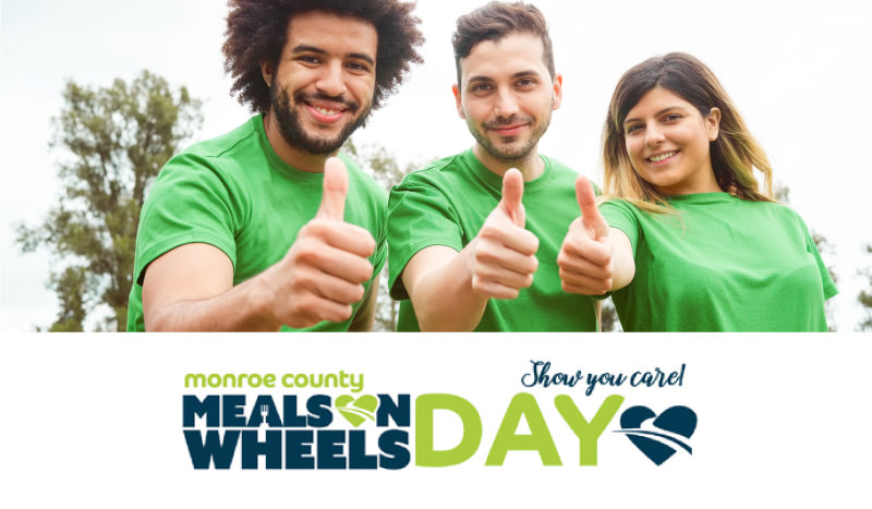 Meals on Wheels day three smiling volunteers giving thumbs up sign while wearing green t-shirts standing outside next to some trees and a pale gray sky