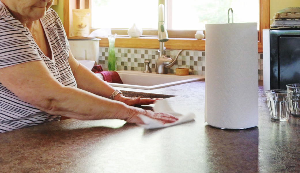 Older woman cleaning kitchen counter with paper towel, roll of paper towels ini holder and glasses also on counter, sink under window in the background