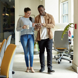 Man and woman dressed casually for dress down day at office, walking through some office chairs with windows in the background