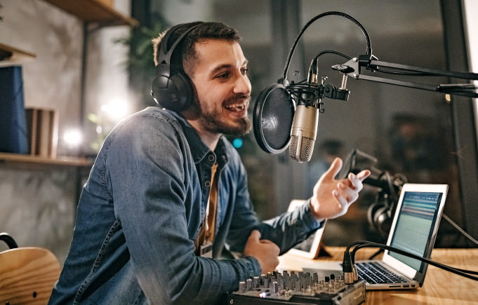 Man wearing a denim button up shirt talking on microphone on radio with laptop on desk