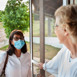 Woman in blue disposable mask visits elderly woman holding open door