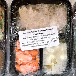 Roasted turkey, gravy, carrots, cauliflower frozen meal in a black tray with plastic wrap over the top and a white label