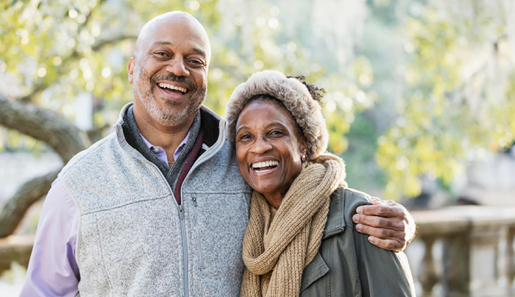 Older couple smiling, wearing winter vest, jacket and fur headband standing outside with sun shining through the trees and a stone fence in the background