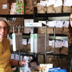 Two women Monroe County Meals on Wheels volunteers at food pantry with shelves of bagged groceries and white binders