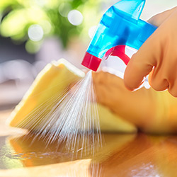 Gloved hand spraying cleaning product from a blue bottle with a yellow cleaning cloth and a wooden table