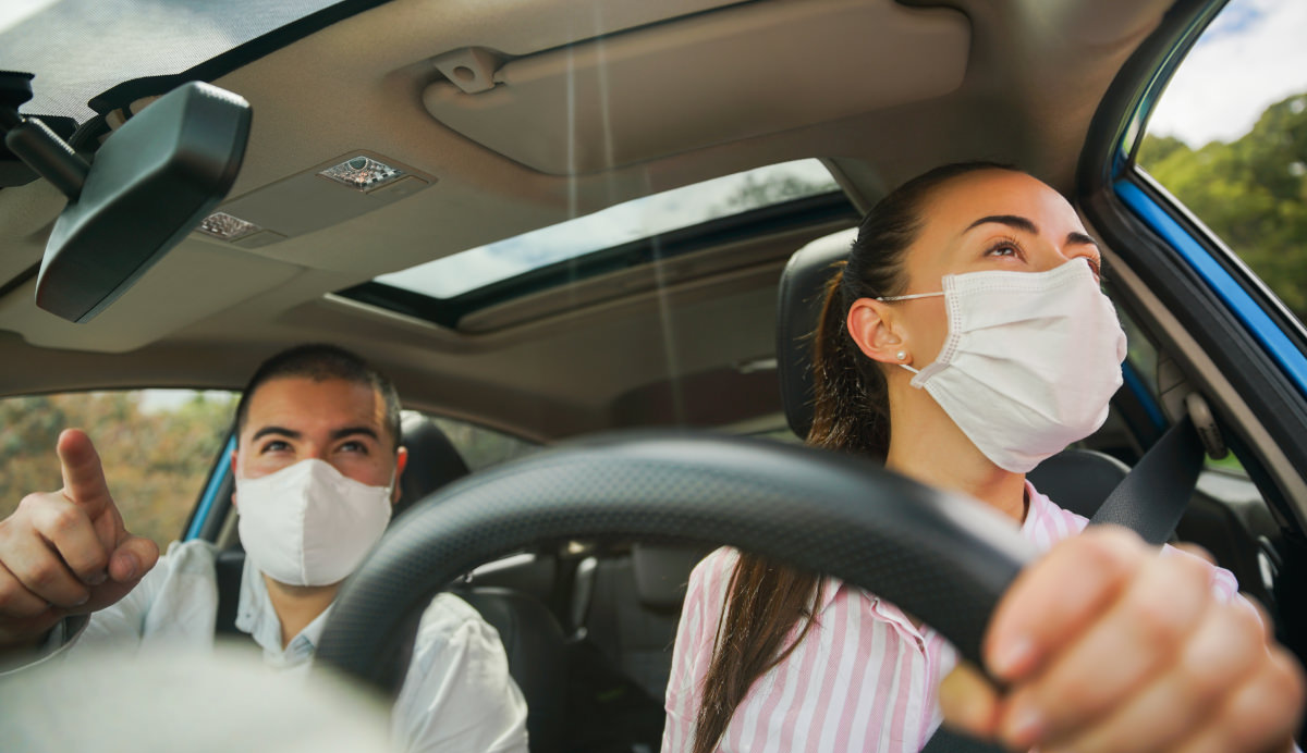 Man and woman in white disposable masks driving a car as seen through the windshield, man is pointing forward