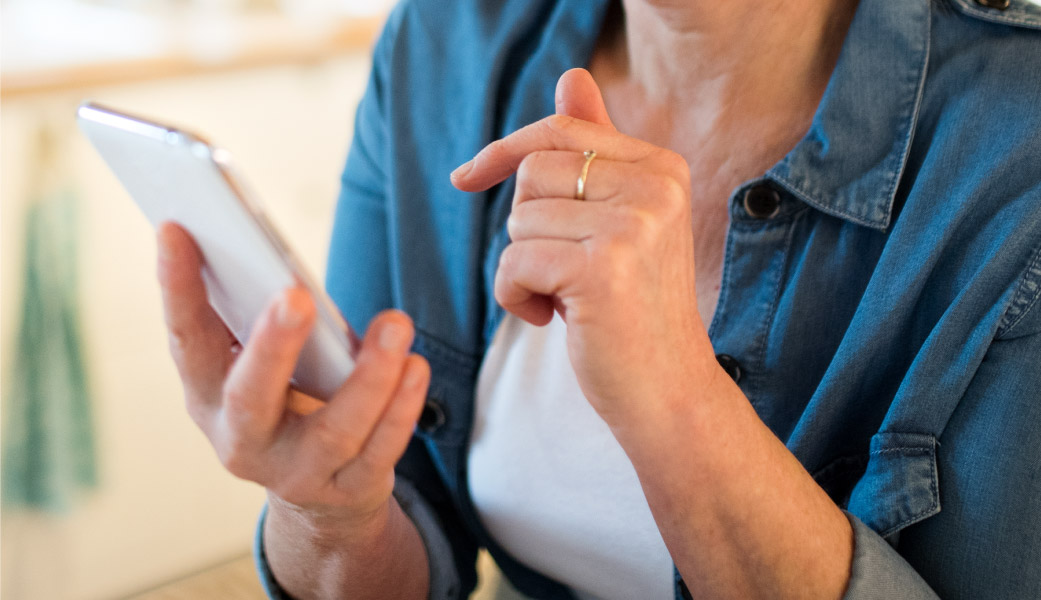 Person wearing a simple gold ring on their hands and holding a white smart phone, also wearing a white shirt and a chambray button up