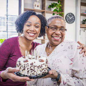 Two women smiling and holding a birthday cake with candles and sprinkles in front of a window and shelves